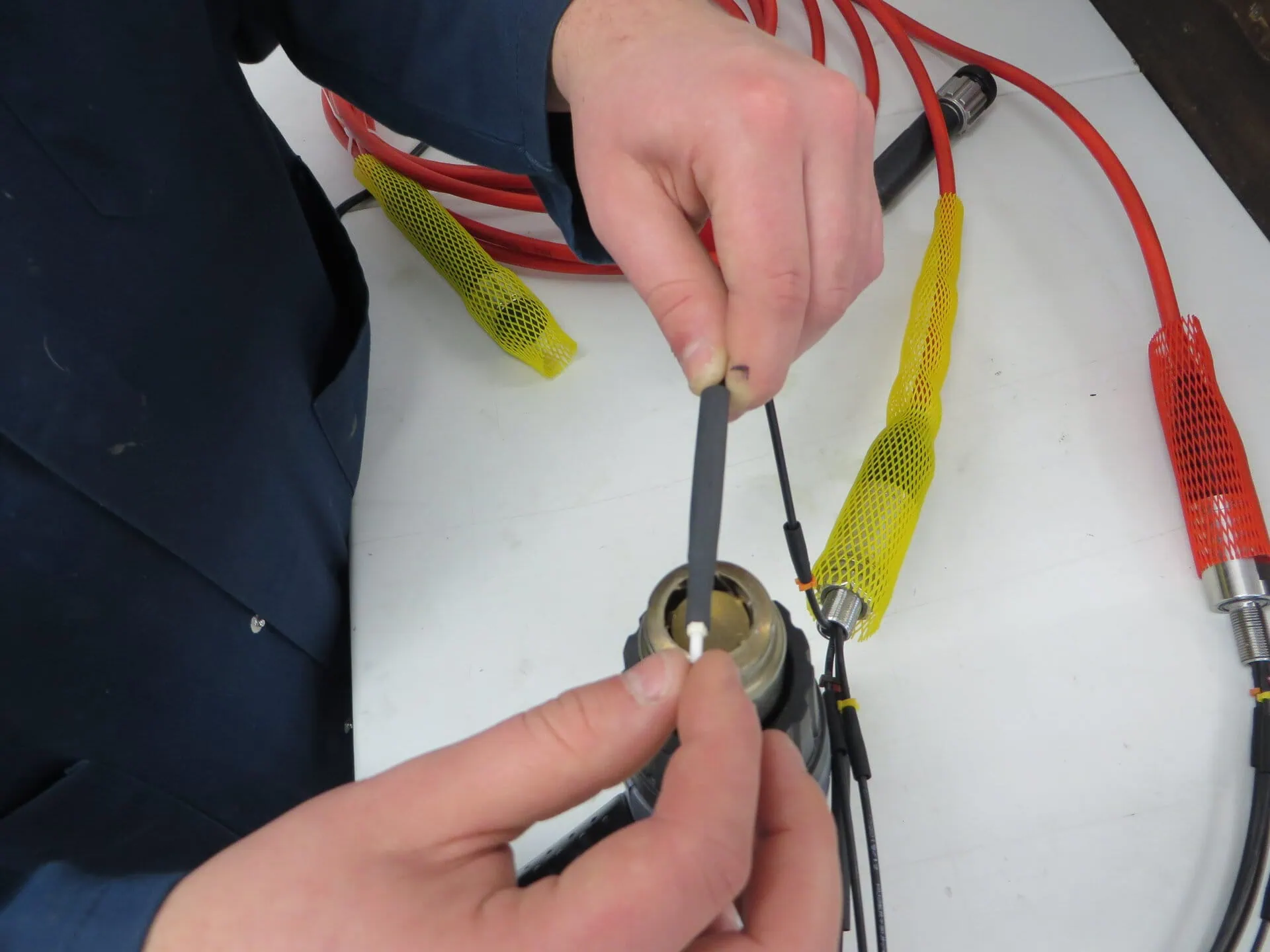 A close-up shot of a person's hands working on a specialized multi-connector cable assembly, applying a piece of black tubing to a white ferrule. The cable has several red and black branches covered in a yellow mesh sheath.