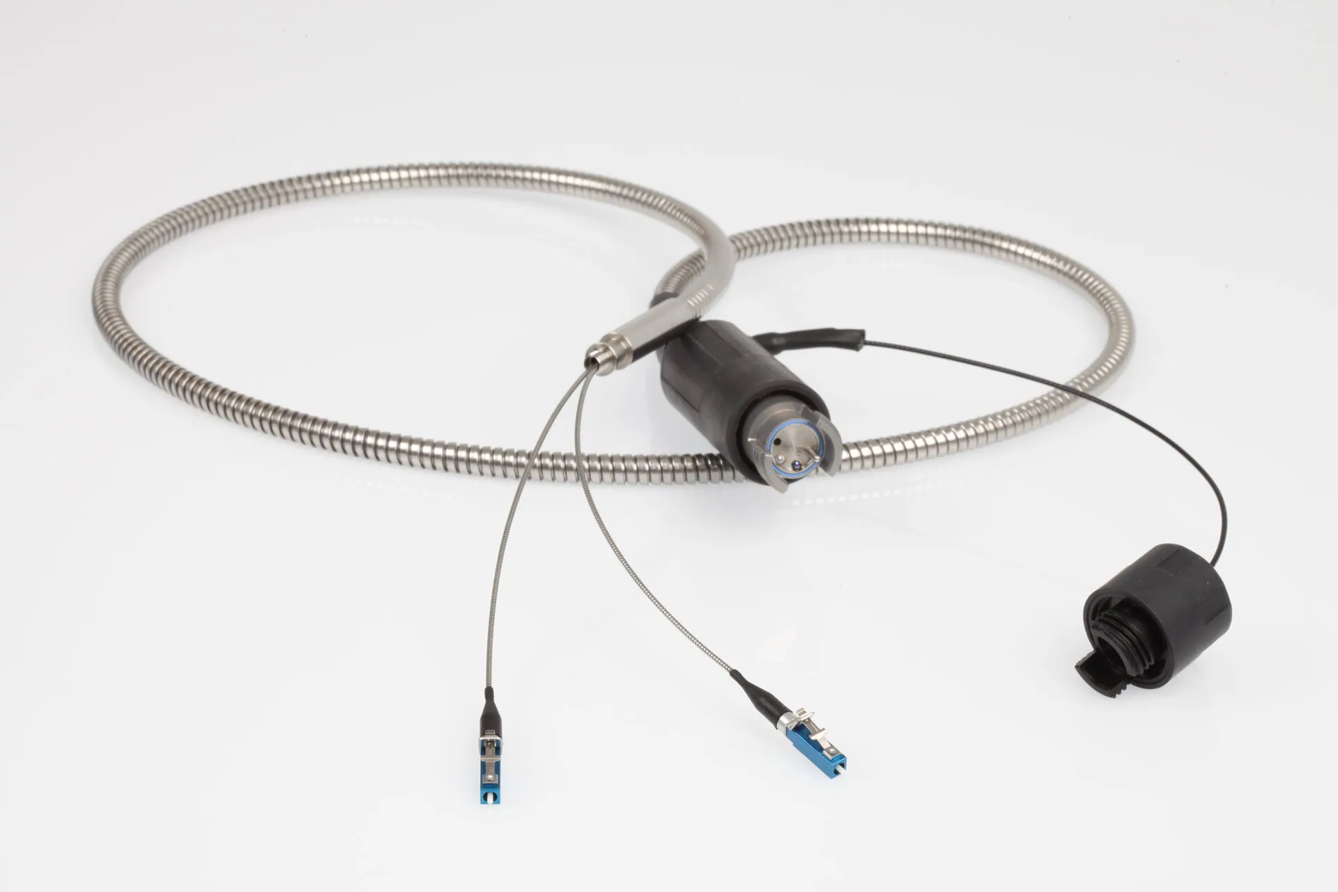 A detailed shot of a coiled, metal-braided fibre optic cable assembly on a white background. A central black component serves as a breakout point, with several smaller cables emerging, two of which terminate in blue LC connectors.