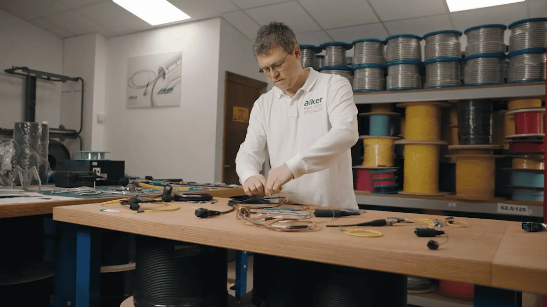 An Alker technician, wearing a white polo shirt with the company logo, is shown assembling a bundle of fibre optic cables and connectors on a workbench in a workshop. Rolls of various cables are visible on shelves in the background.