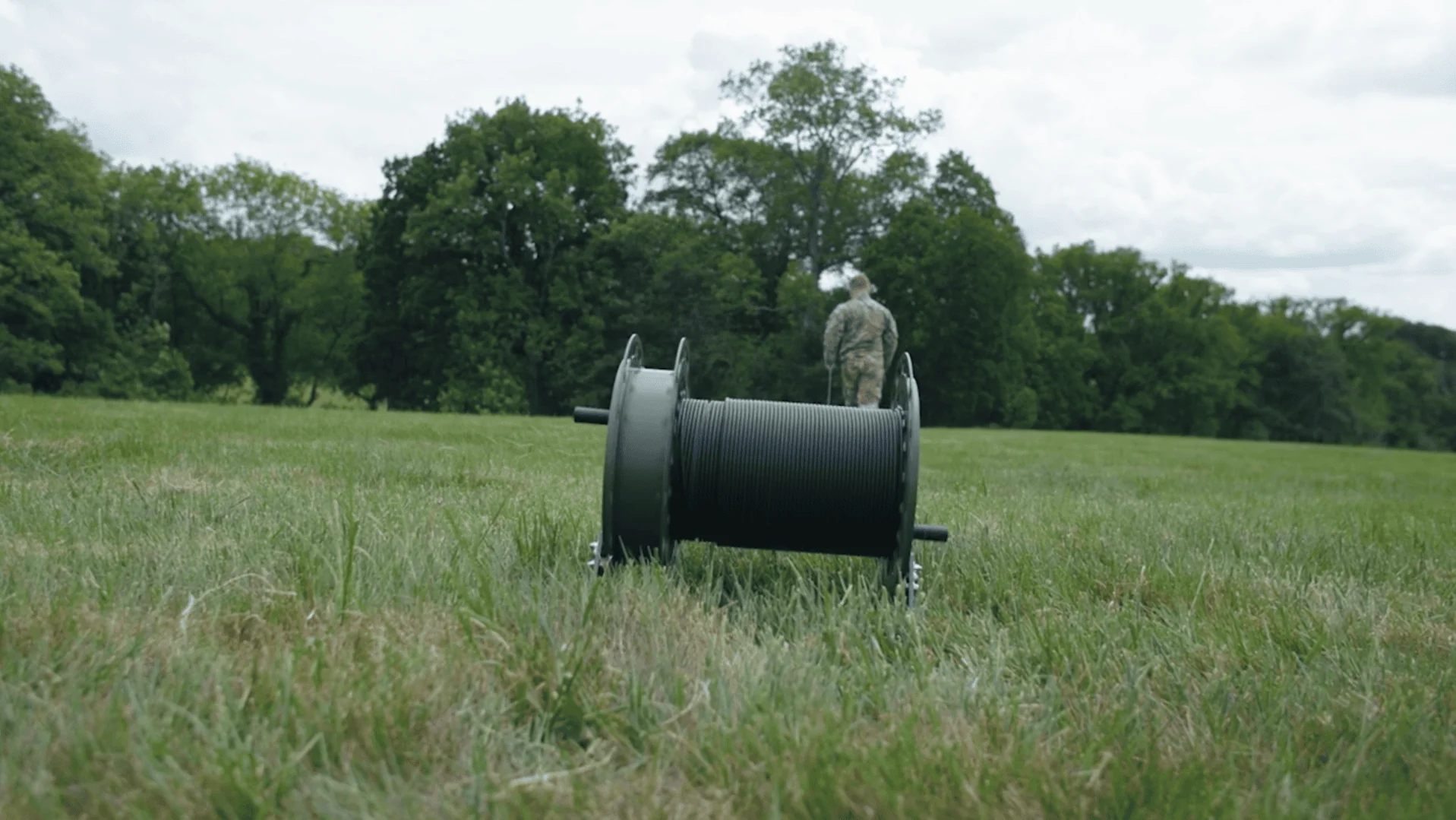 An Alker employee, wearing a company polo shirt, smiles while holding the hinged side cover of a large, olive-green deployable fibre optic cable reel on a workbench.