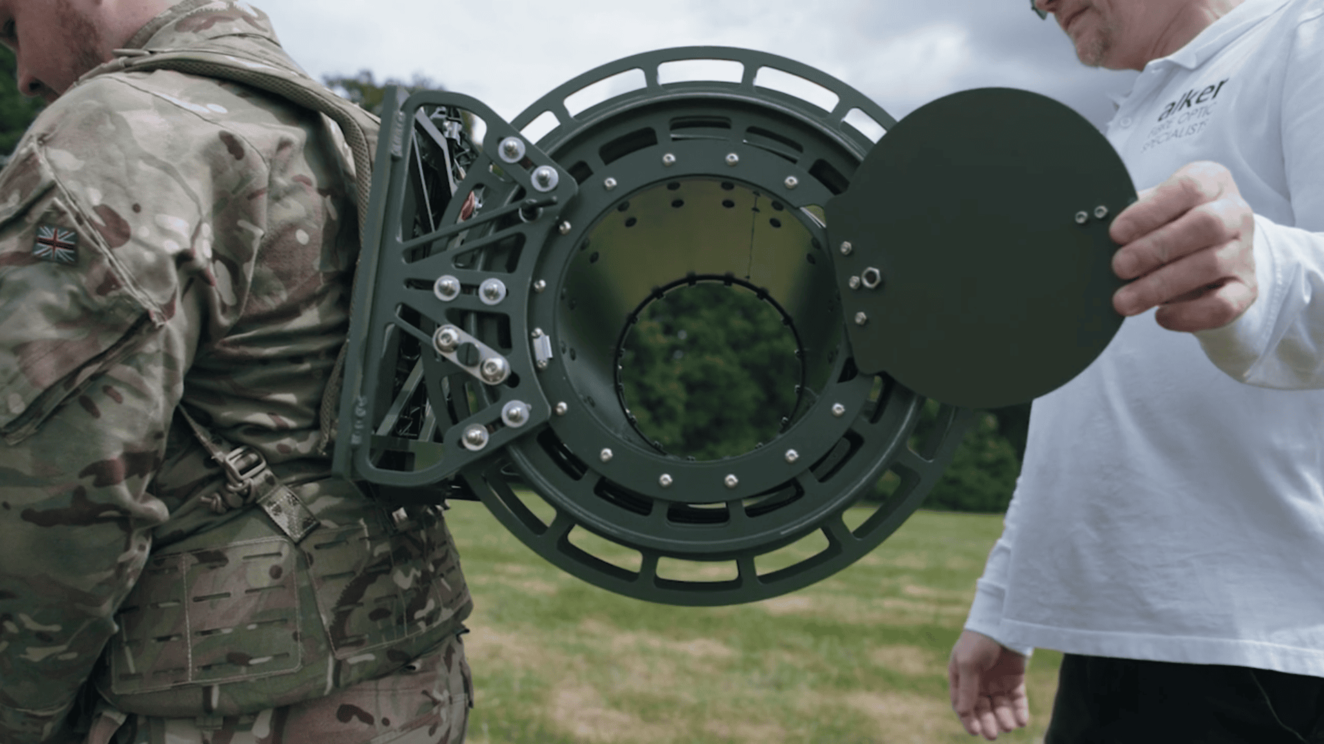 An Alker employee in a white polo shirt holds the cover for a tactical fibre optic cable dispenser, which is worn as a backpack by a soldier in a camouflage uniform, demonstrating the equipment in a grassy field.