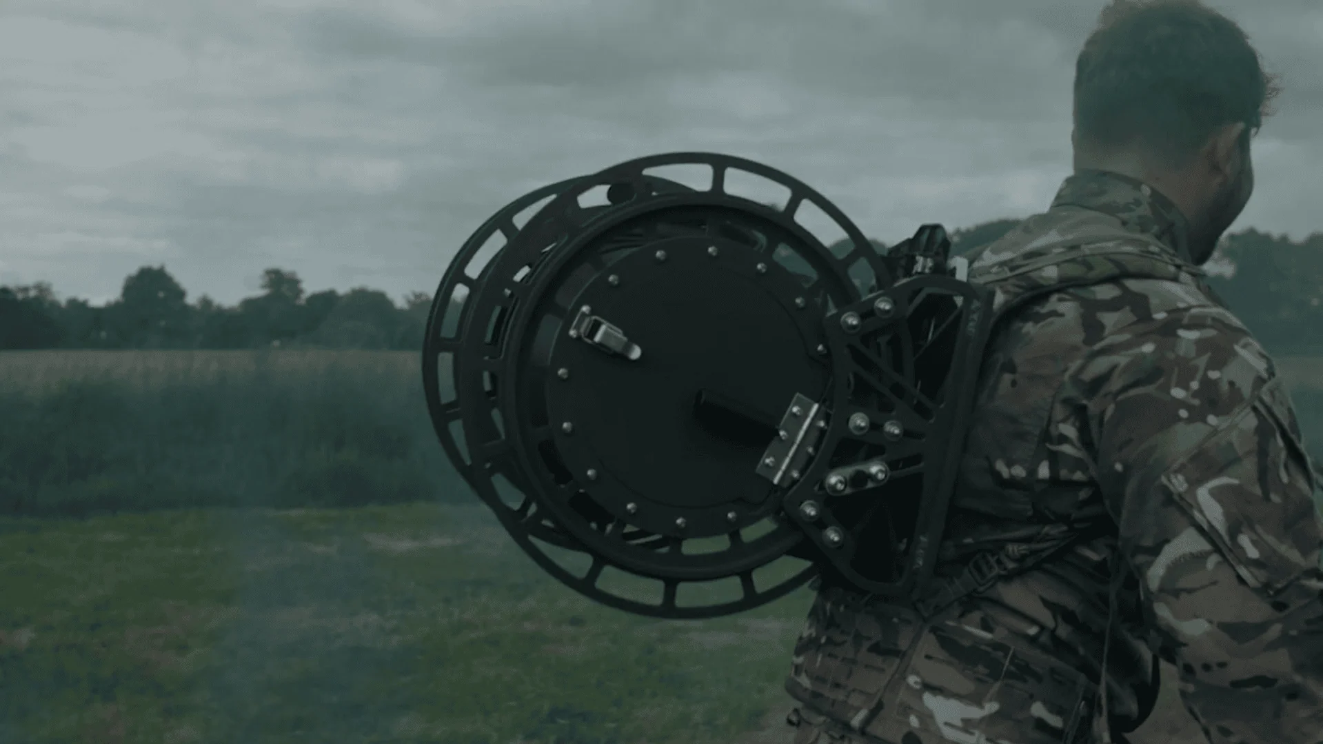 A person in a camouflage uniform, viewed from the back, is walking through a field while carrying a large, specialized black tactical fibre optic cable dispenser on their back.