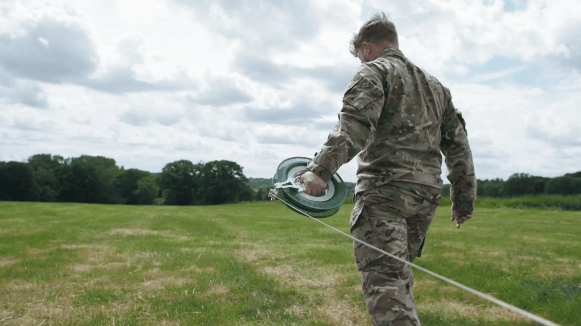 A soldier in a camouflage uniform, seen from behind, walks through a grassy field while unwinding a fibre optic cable from a small, handheld reel.