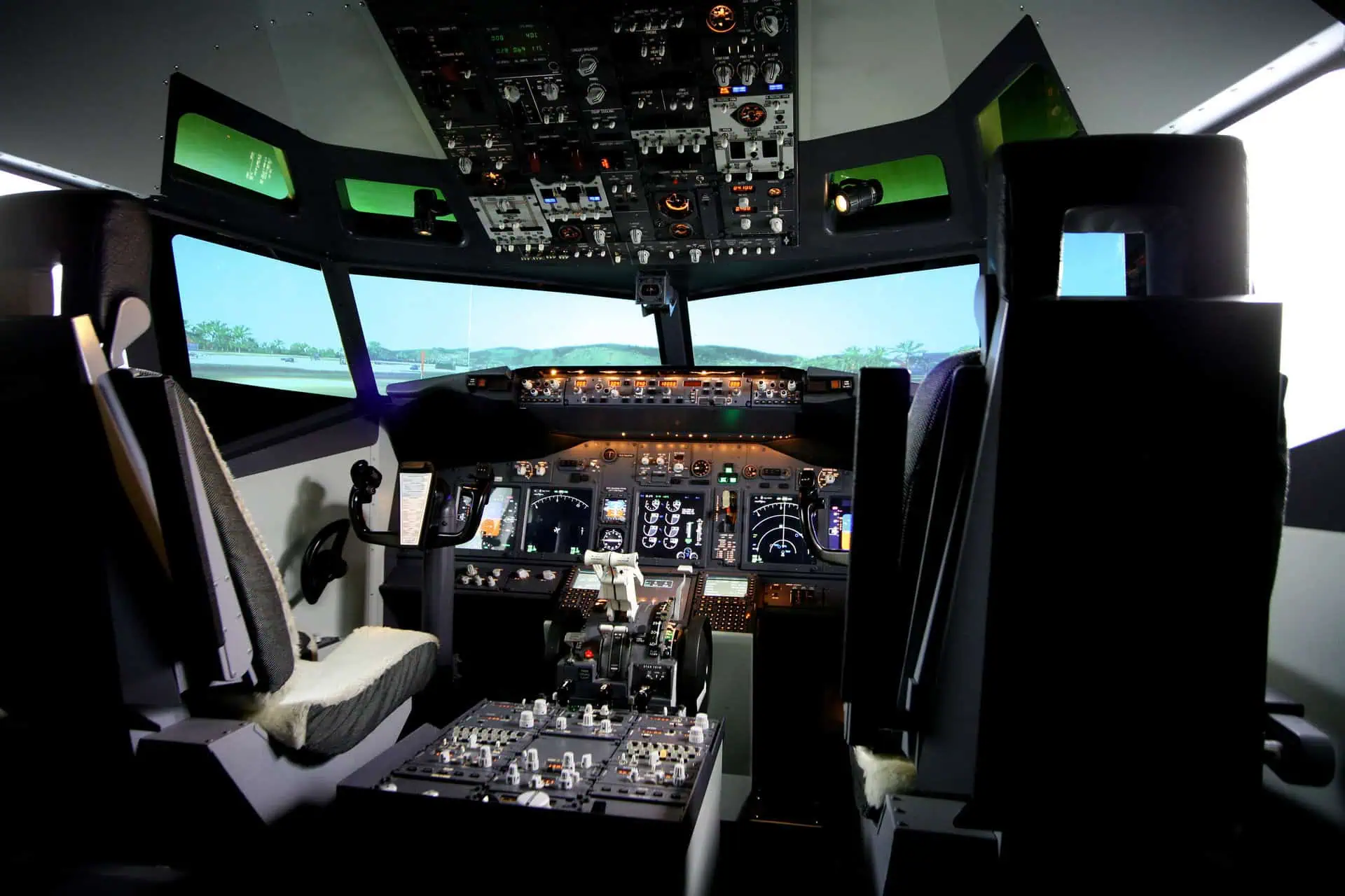 A detailed view from inside the cockpit of a modern commercial aircraft flight simulator, showing the complex array of instruments, controls, and pilot seats, with a simulated airfield visible through the windows.