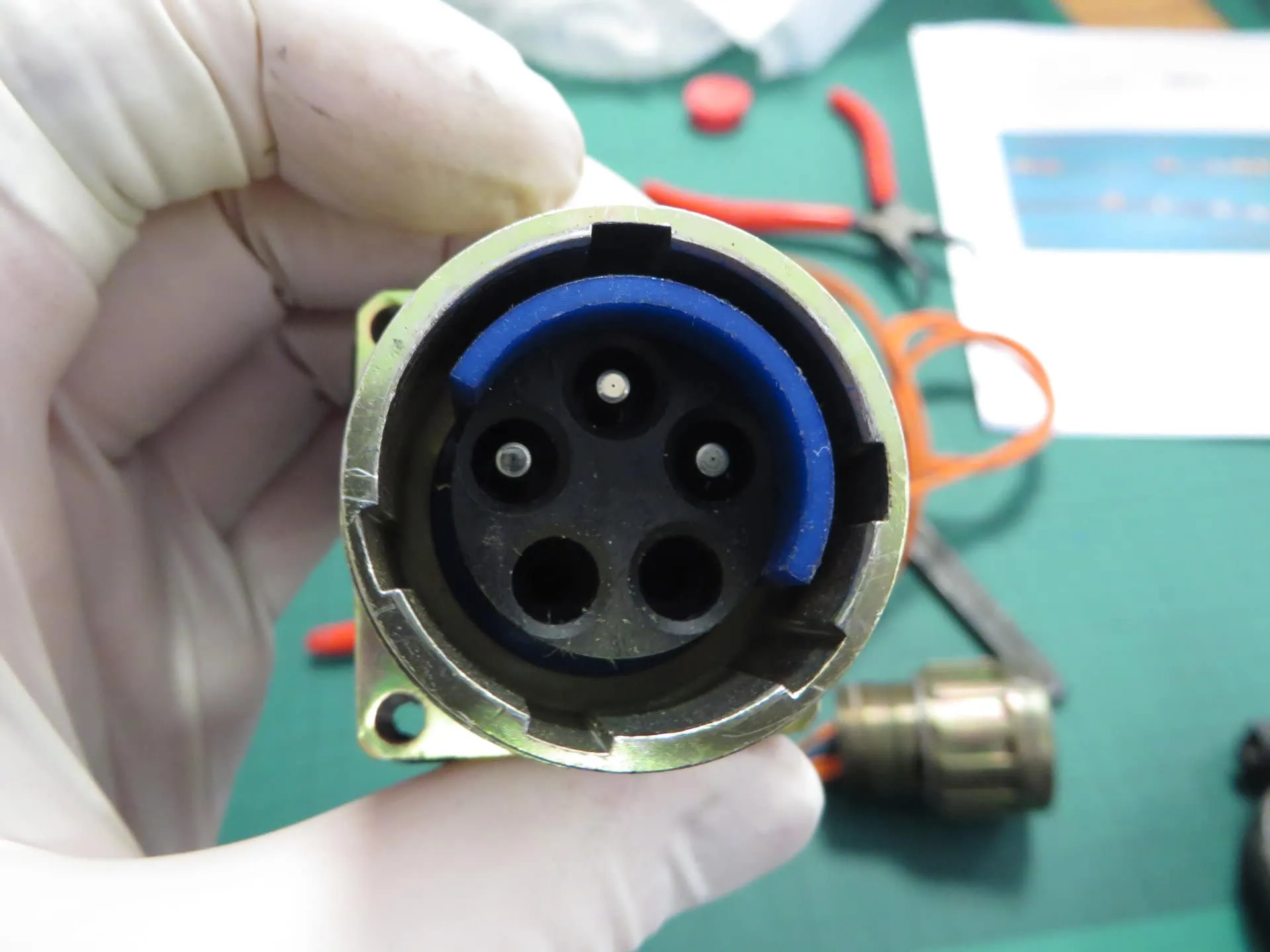 A gloved hand holds a circular metal connector socket with a black plastic core, a large blue ring, three metal pins, and three empty ports visible inside.