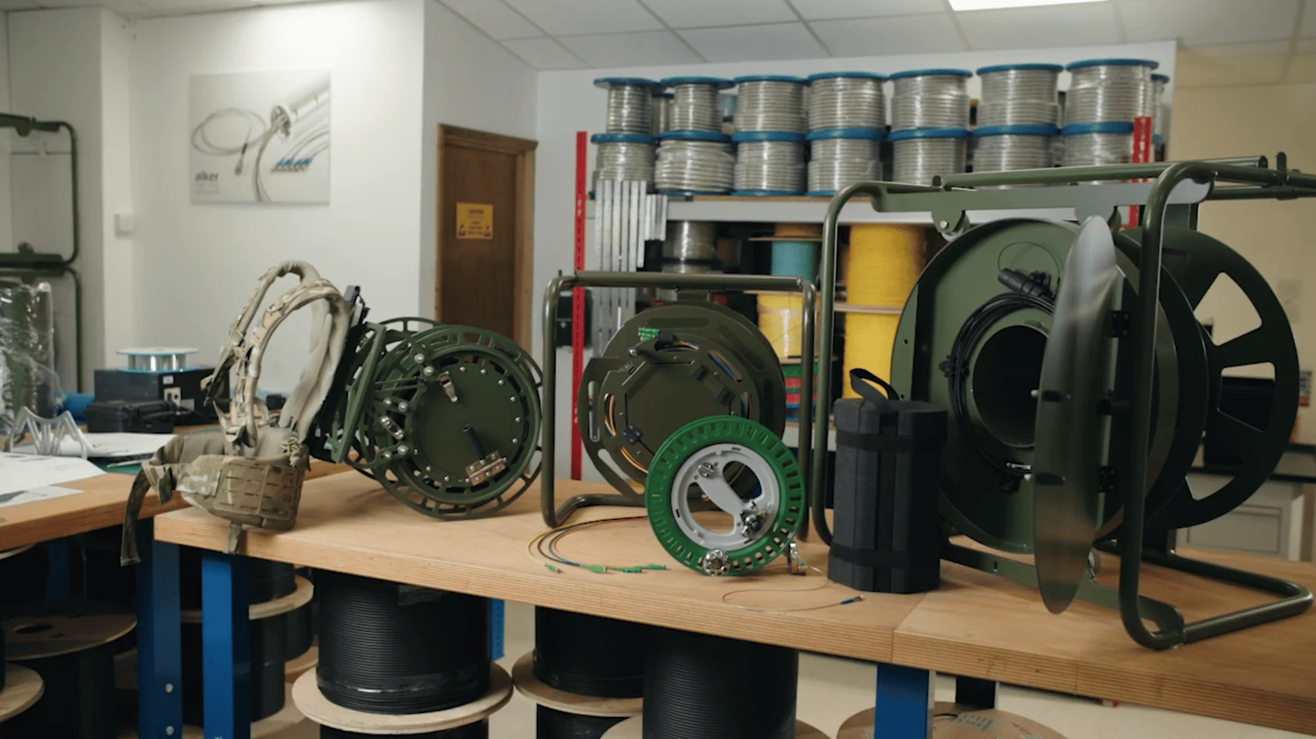 A collection of military and field-grade fibre optic equipment is arranged on a workbench, including several tactical cable dispensers of varying sizes, a repair kit, and other specialized accessories.