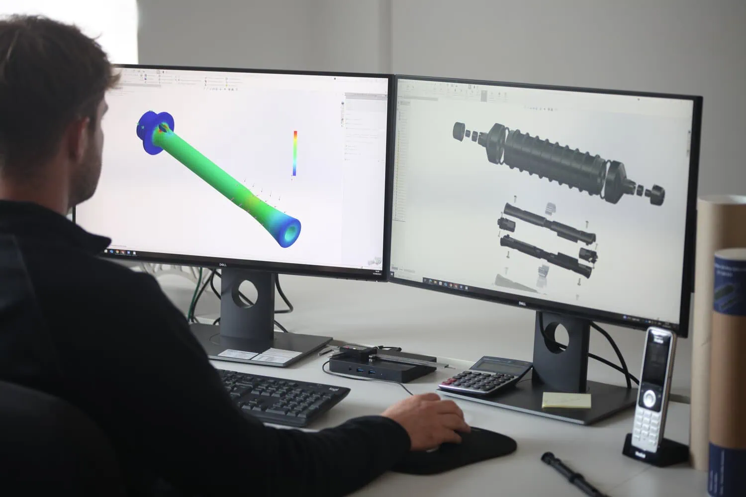 A person is shown from behind, sitting at a desk with two computer monitors. The screens display 3D computer-aided design (CAD) models of fibre optic components, including stress analysis and an exploded view of a ruggedized cable connector.
